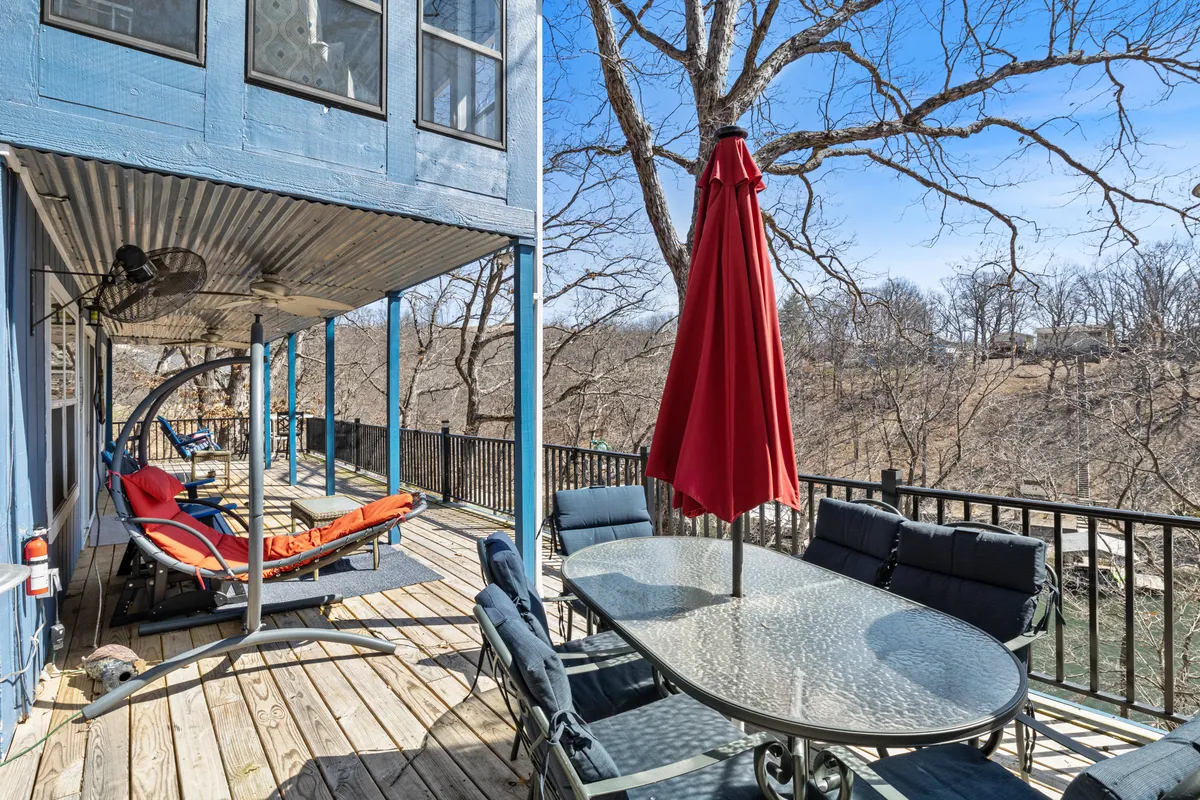 An inviting outdoor deck scene featuring a red umbrella shading a glass dining table, hammock chair, and wooden deck flooring. The area overlooks water, creating the perfect spot for alfresco dining and relaxed gatherings.