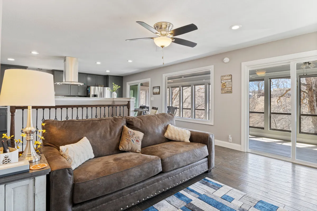A cozy living room featuring a comfortable sofa, a large rug, windows that let in natural light, a ceiling fan, and a view of the kitchen area. The space feels inviting and is well-suited for family gatherings or relaxing evenings.