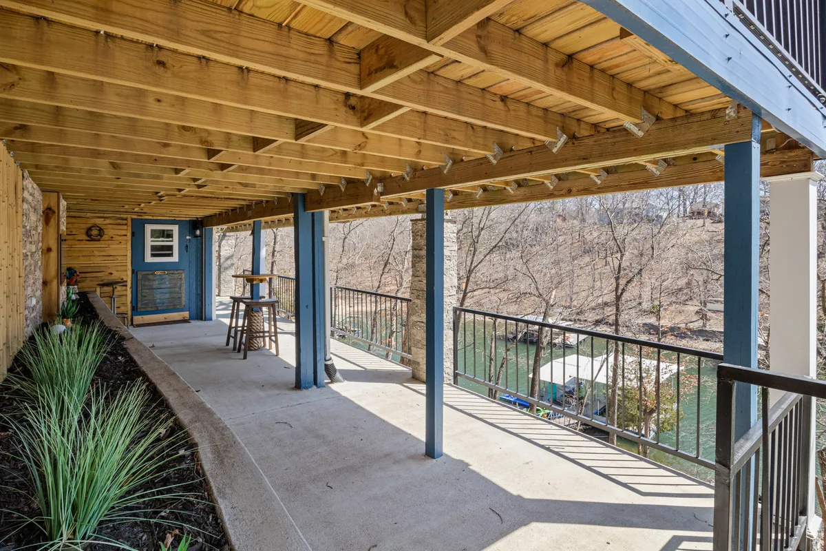 An inviting view of a covered porch area with wooden pillars and a bar stool, looking out over the river. The concrete walkway and railing suggest easy access for gatherings, while the scenic water view sets a serene backdrop for morning or evening activities. The atmosphere is tranquil, ideal for both intimate chats and group celebrations.