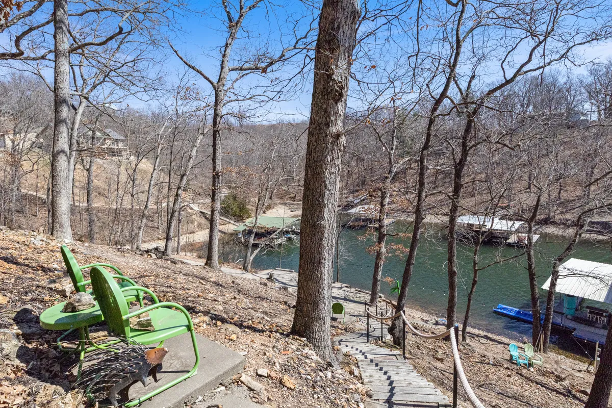 This image showcases a tranquil exterior view with green chairs arranged on a porch or deck, steps leading down to the water, and a picturesque river scene in the background. The mood is peaceful, suggesting opportunities for relaxing by the water, enjoying morning coffee, or winding down as the sun sets. Hints of bonfire gatherings and water-based activities are present in the scene.
