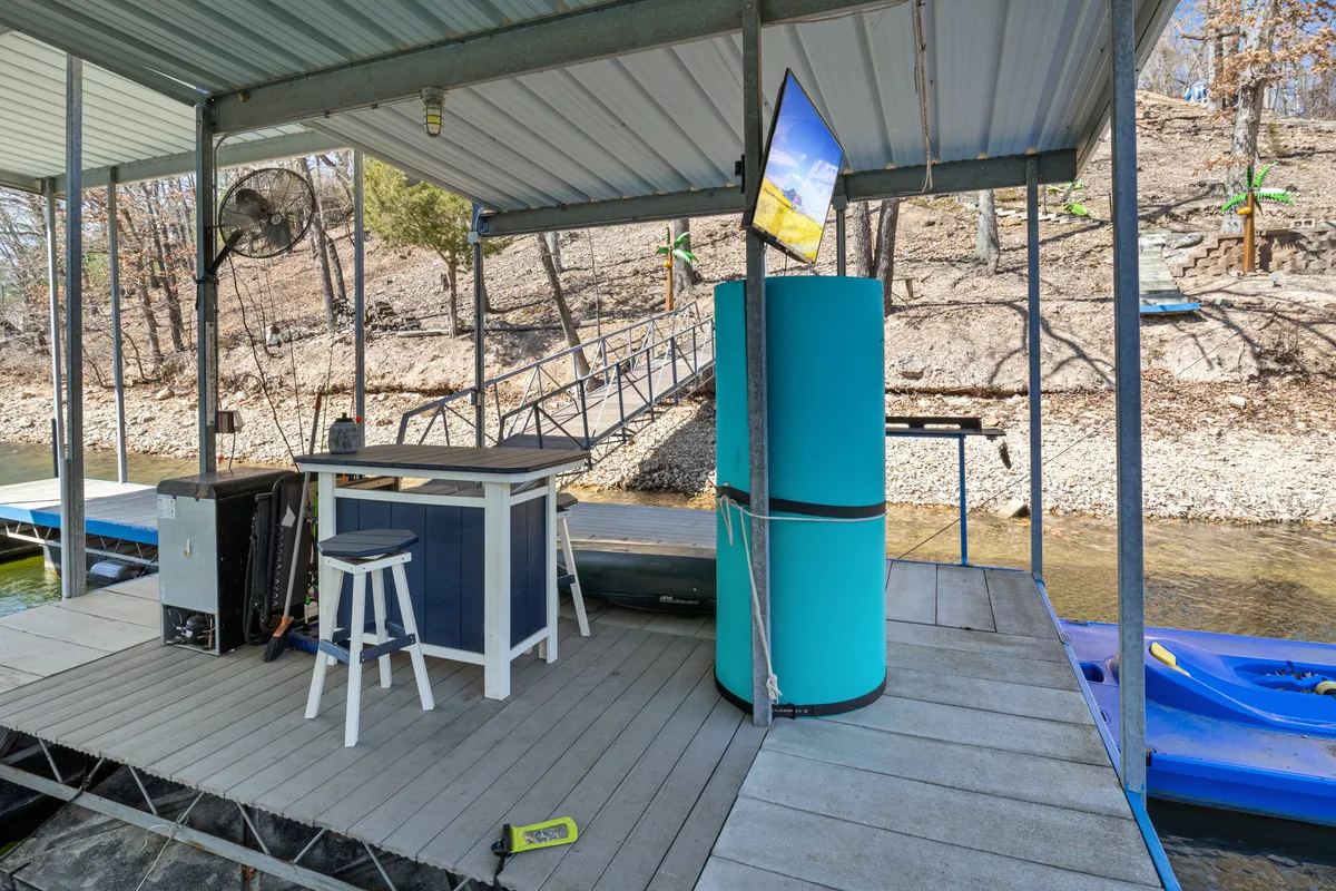 This exterior image features a wooden dock extending over the water, complemented by an outdoor bar area with a blue cooler and a television mounted on the wall. The scene is set for lakeside gatherings, fishing or kayaking right from the dock, and enjoying the tranquility of the lake. The space is inviting for social or family activities, and the dock is large enough for both relaxing and launching watercraft.