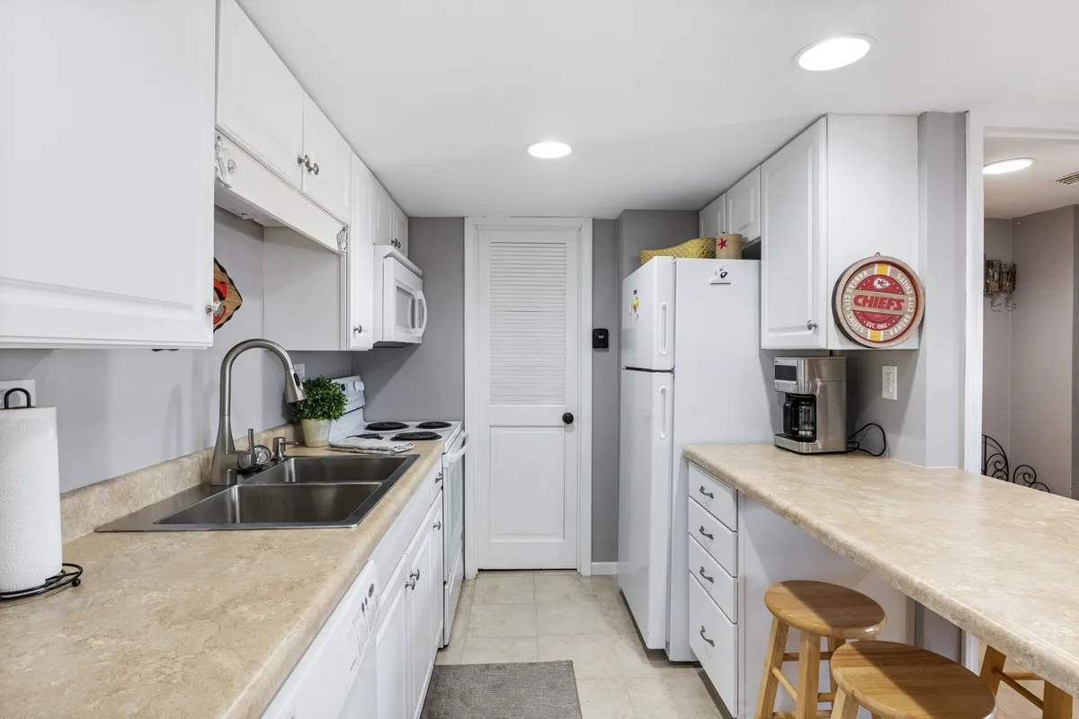 A cozy, functional kitchen area with a long countertop, stools for casual seating, white cabinetry, and a variety of appliances. The layout encourages multiple cooks or informal family gatherings.