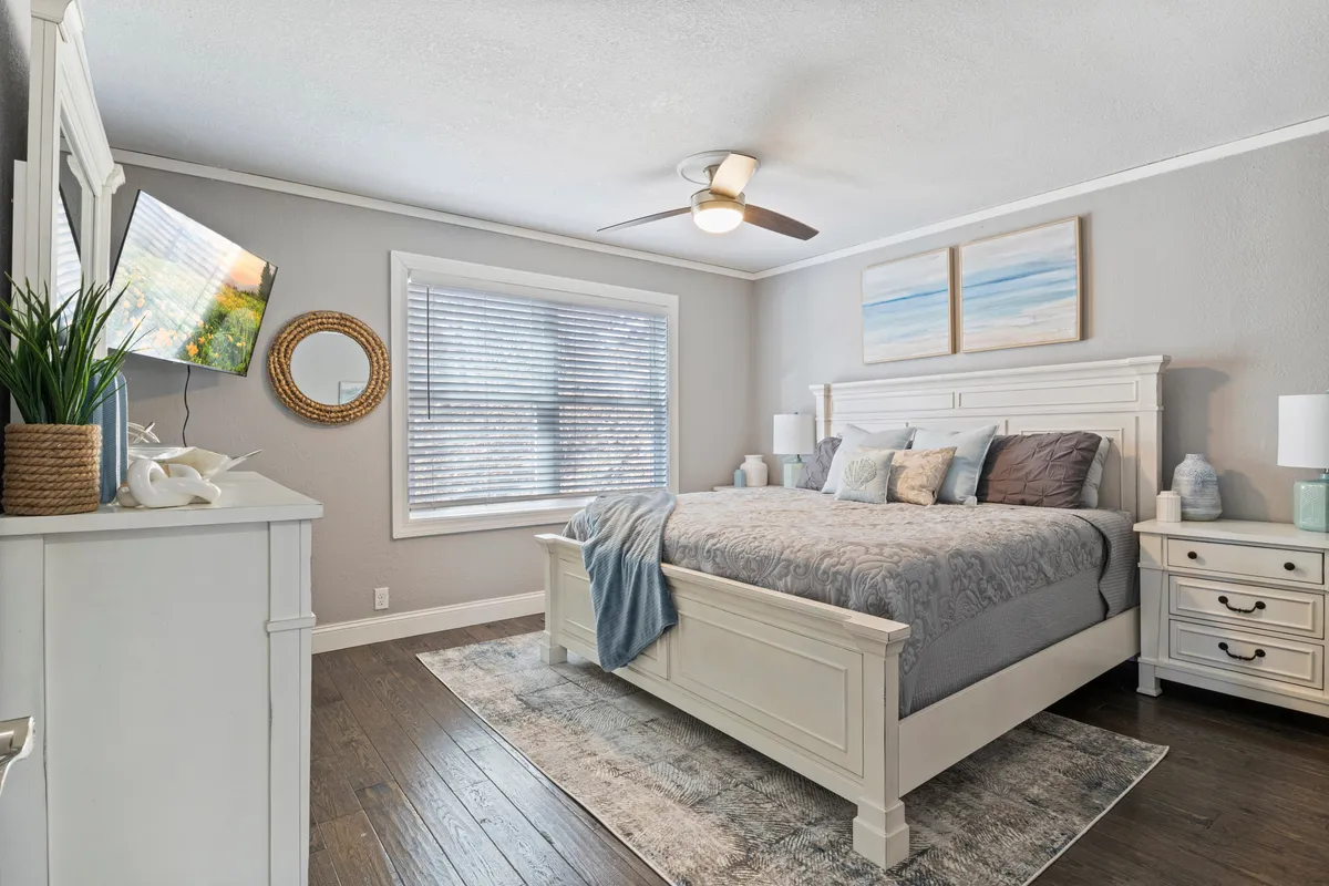 A serene bedroom featuring a comfortable bed adorned with decorative pillows, tasteful artwork on the wall, a flat-screen TV, and a window with blinds allowing natural light to softly illuminate the space. The overall decor is cozy and inviting, ideal for relaxation and restful sleep.